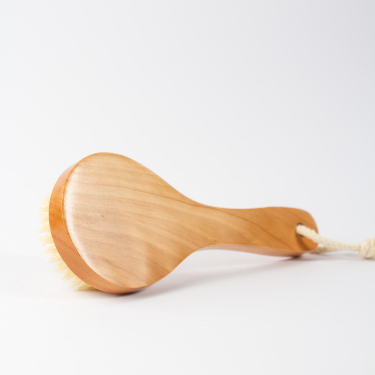 A wooden boar brush with a light natural wood head and handle, cream-colored bristles, and a white hanging cord, photographed on a clean white background.