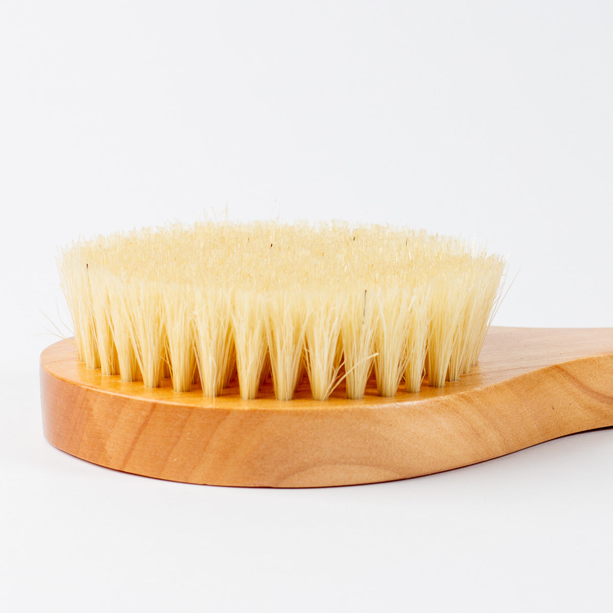 A close-up of a boar bristle body brush with a light natural wood handle and dense cream-colored bristles, set against a plain white background.