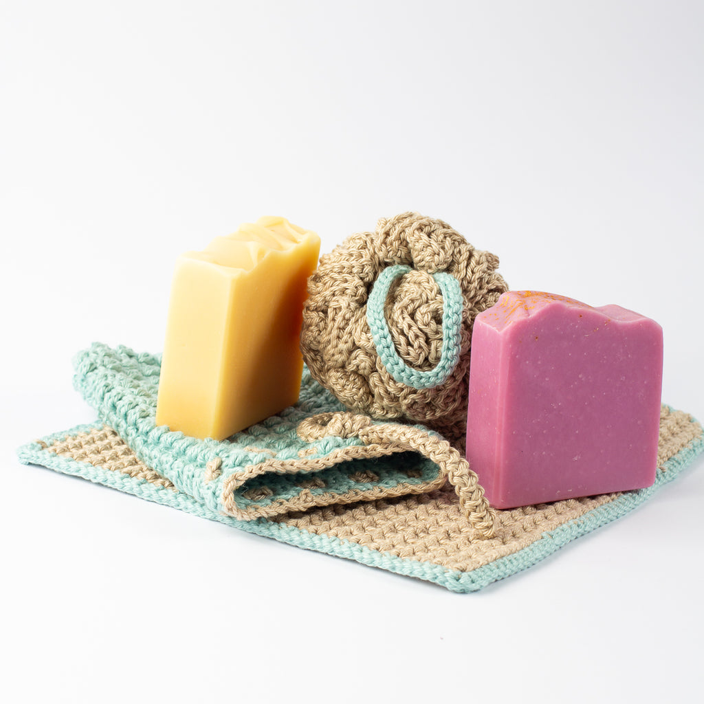 A handmade crochet bath and shower accessory set with washcloth, shower pouffe or sponge and soap saver bag in beige and sage green, styled with 2 bars of handmade soap, on a clean white background.