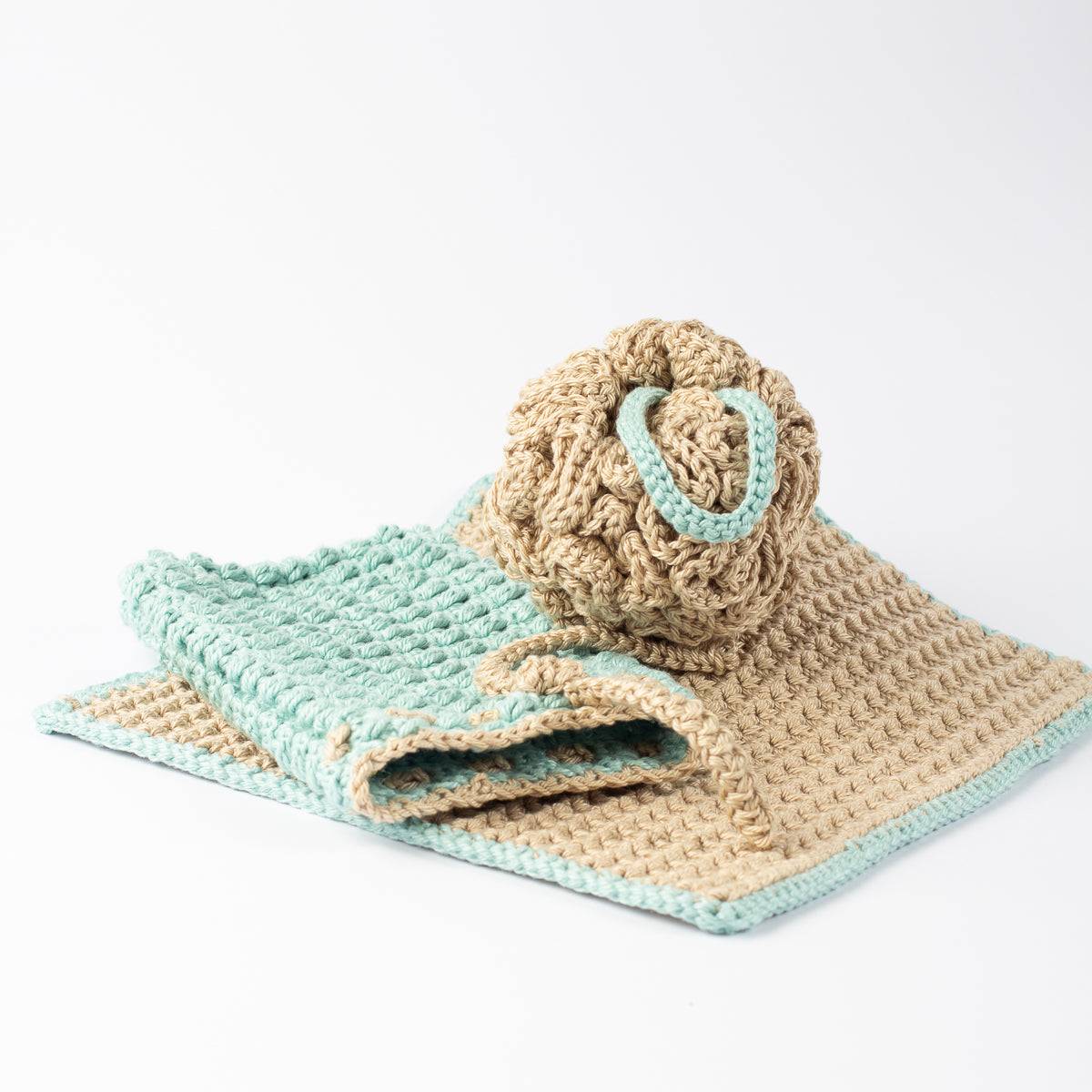 A handmade crochet bath and shower accessory set with washcloth, shower pouffe or sponge and soap saver bag in beige and sage green, on a clean white background.