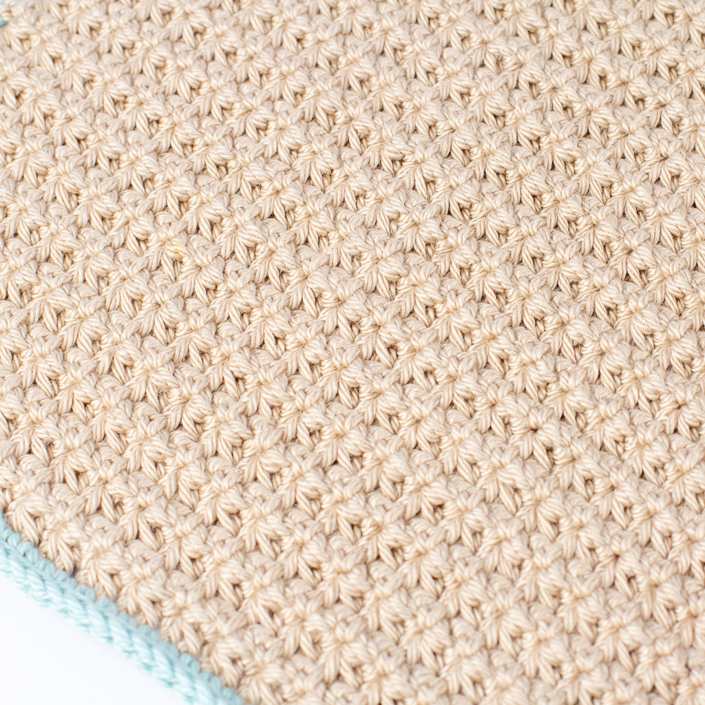 Close-up of a textured crochet washcloth in a warm beige color, showing a detailed starburst or rosette stitch pattern. A small portion of a light sage green knitted edge is visible in the bottom-left corner against a white background.