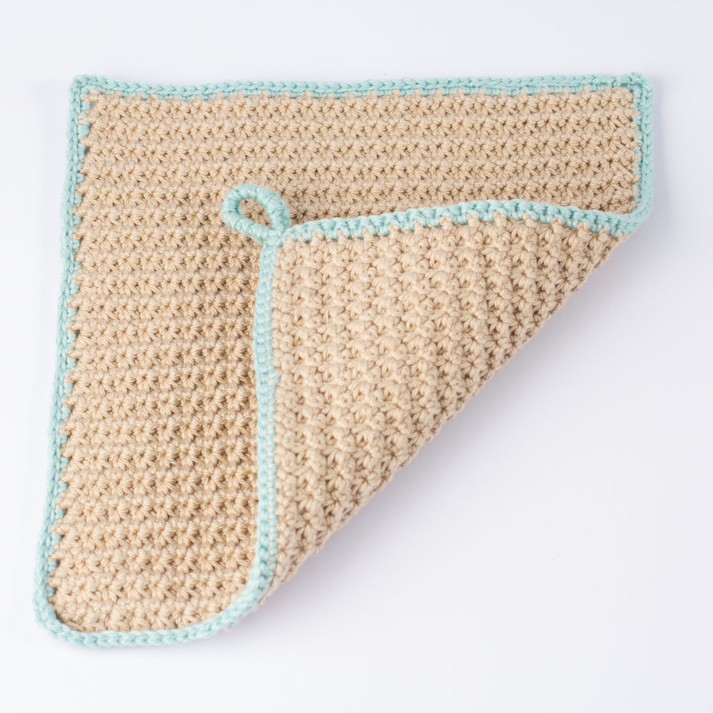 A square crocheted cloth in a warm beige/tan color with a light sage green border, shown on a plain white background with one corner folded over to reveal the reverse side.