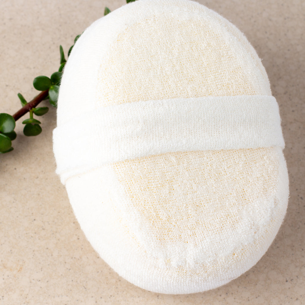ECONIQUE Natural Loofah Body Sponge reverse side showing soft white fabric encasing and hand strap, resting on a stone surface alongside a small succulent branch