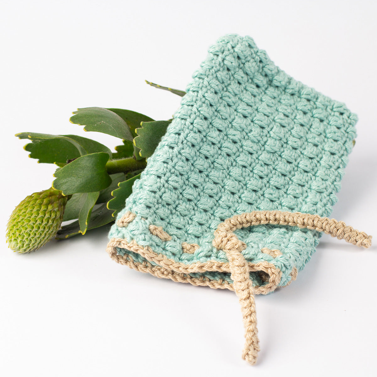 ECONIQUE sage green cotton soap saver pouch laid with South African fynbos botanical sprig on white surface